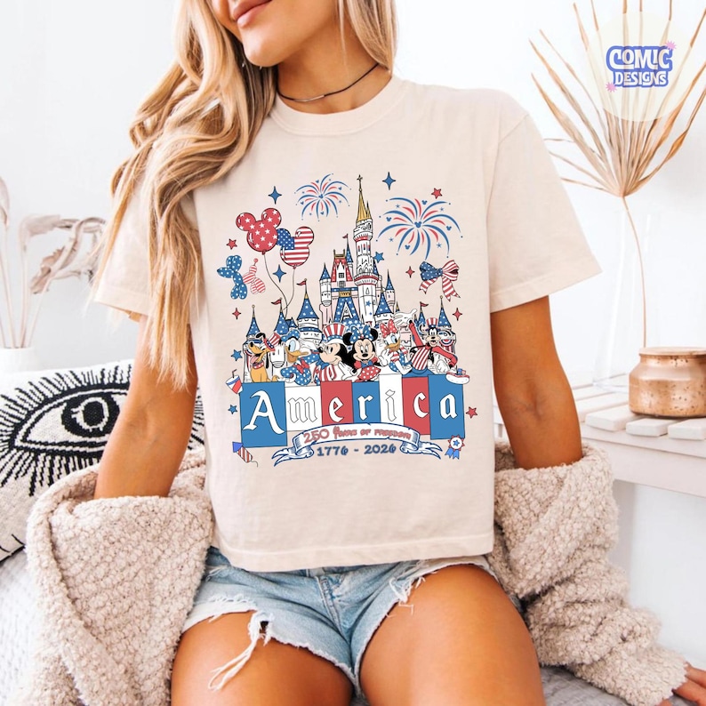 May include: A cream-colored t-shirt featuring a graphic print of a castle, fireworks, and cartoon characters wearing patriotic red, white, and blue clothing. The text reads America, 250 Years of Freedom, and 1776-2026 in a decorative banner.