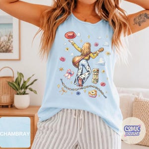 May include: A light blue sleeveless tank top featuring a graphic illustration of a person with long hair holding a guitar, wearing a yellow top and cow-print pants. The design includes a red hat, flowers, stars, clouds, and blocks with the letters T, S, and S.