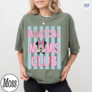 K&ouml;nnte beinhalten: Olivgr&uuml;nes T-Shirt mit dem Text "MAGICAL MOMS CLUB" in Pink, mit einer Minnie-Maus-Grafik und einer Schloss-Illustration. Das Design ist vor einem Hintergrund aus hellblauen vertikalen Streifen angeordnet.