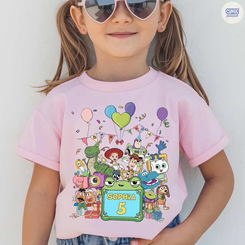 May include: A light pink short-sleeve t-shirt featuring a colorful graphic of Toy Story characters, balloons, and birthday bunting. A central green frame displays the name Sophia and the number 5 in yellow text, surrounded by gift boxes.
