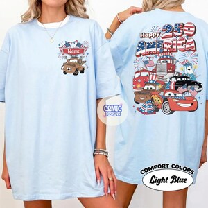 May include: Pale blue t-shirt with a cartoon design of a brown truck and the word "Name" above it. The back of the shirt features the words "Happy 250 America" and various cartoon vehicles. The shirt is labeled "Comfort Colors Light Blue."