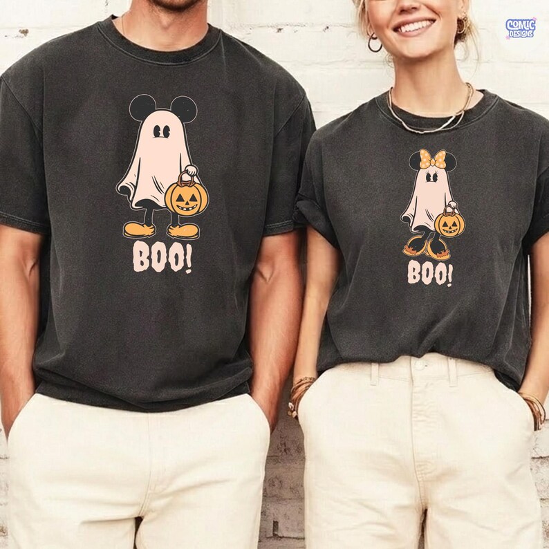 May include: Two dark gray t-shirts featuring a cartoon ghost with Mickey Mouse ears holding a jack-o'-lantern. The ghost is wearing yellow shoes and the word "BOO!" is printed below. The shirts are a Halloween theme.