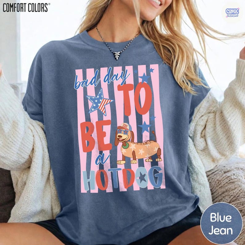May include: Blue jean Comfort Colors t-shirt with the phrase "bad day to be a hotdog" in a playful font. The design includes a cartoon dachshund dressed as a hotdog, with stars and stripes.