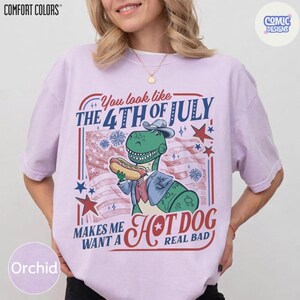 May include: Orchid Comfort Colors t-shirt with a graphic featuring a green dinosaur in a suit holding a hot dog in front of an American flag. Text reads "You look like the 4th of July makes me want a hot dog real bad."