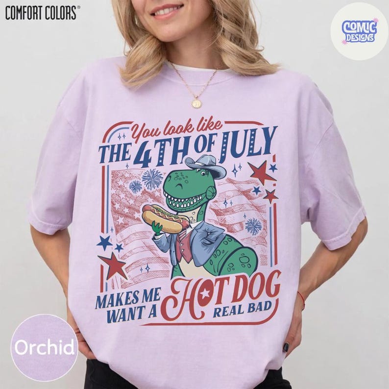 May include: Orchid Comfort Colors t-shirt with a graphic featuring a green dinosaur in a suit holding a hot dog in front of an American flag. Text reads "You look like the 4th of July makes me want a hot dog real bad."
