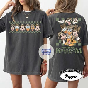 Pode incluir: Camiseta cinza escuro com personagens da Disney usando chap&eacute;us de safari. A frente mostra uma fileira de personagens, a parte de tr&aacute;s um grupo com "Animal Kingdom" e "Est. 1998". A camiseta &eacute; rotulada como "Comfort Colors Pepper".