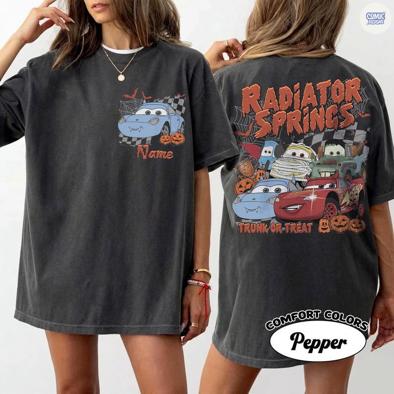 May include: A charcoal grey t-shirt featuring a small graphic on the front chest of a blue car with fangs and pumpkins. The back displays a large graphic with the text Radiator Springs and Trunk or Treat, featuring multiple cars in costumes.