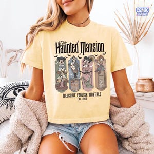 May include: Yellow t-shirt featuring "The Haunted Mansion" graphic. The design includes cartoon characters and the text "WELCOME FOOLISH MORTALS Est. 1969". The shirt has short sleeves and a crew neck.