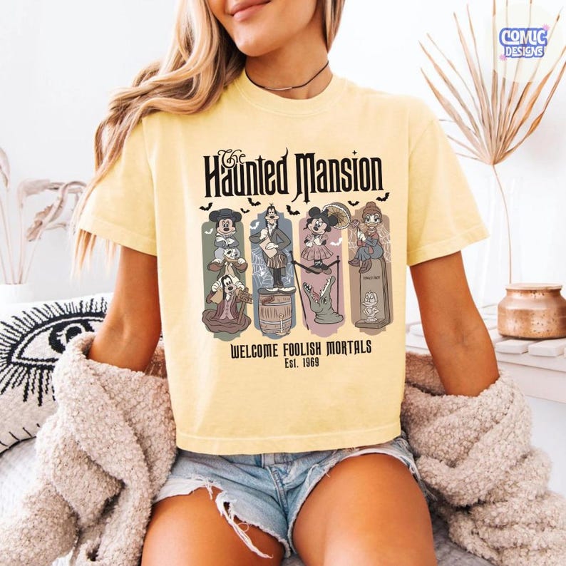 May include: Yellow t-shirt featuring "The Haunted Mansion" graphic. The design includes cartoon characters and the text "WELCOME FOOLISH MORTALS Est. 1969". The shirt has short sleeves and a crew neck.