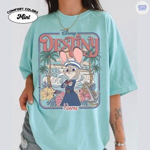 Op de afbeelding: Mintkleurig T-shirt met een Disney "Destiny"-afbeelding. Het ontwerp bevat een cartoonkonijn in een matrozenoutfit, palmbomen en een boot. Het shirt heeft de woorden "Comfort Colors" en "Mint" erop gedrukt.