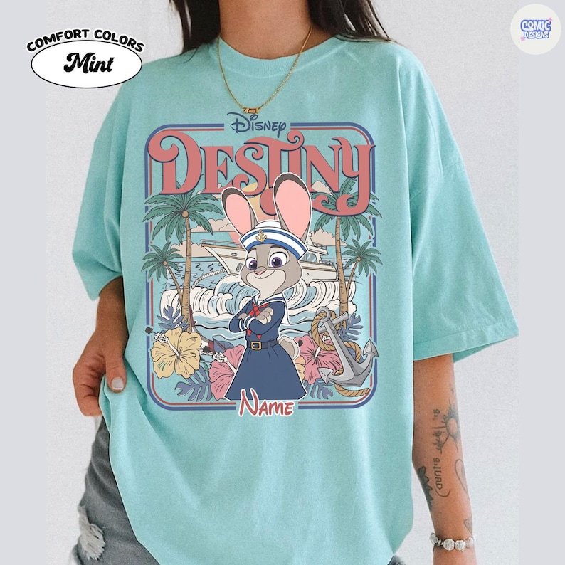 Op de afbeelding: Mintkleurig T-shirt met een Disney "Destiny"-afbeelding. Het ontwerp bevat een cartoonkonijn in een matrozenoutfit, palmbomen en een boot. Het shirt heeft de woorden "Comfort Colors" en "Mint" erop gedrukt.
