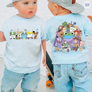 May include: A light blue t-shirt featuring the name HENRY in colorful character-themed letters on the front and a graphic print of various animated toy characters gathered around a purple bag on the back, paired with light blue denim jeans.