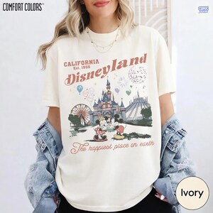 Op de afbeelding: Ivoor kleurig T-shirt met een Disneyland afbeelding. Het ontwerp bevat het Disneyland logo, een kasteel, Mickey en Minnie Mouse, en de tekst "The happiest place on earth". Het shirt is van het merk Comfort Colors.