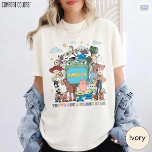 May include: An ivory t-shirt featuring a colorful graphic of various animated toy characters surrounding a central green frame with the name AMELIA printed inside. Below the characters, the text reads YOU'VE GOT A FRIEND IN ME in multicolored letters.