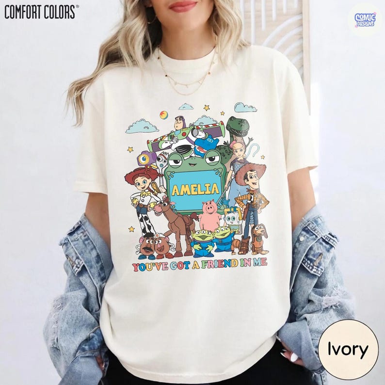 May include: An ivory t-shirt featuring a colorful graphic of various animated toy characters surrounding a central green frame with the name AMELIA printed inside. Below the characters, the text reads YOU'VE GOT A FRIEND IN ME in multicolored letters.