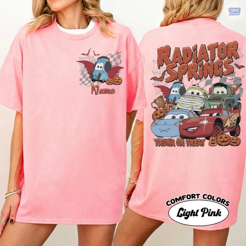May include: A light pink t-shirt featuring a small graphic on the front left chest of a blue car character with bat wings and pumpkins, with the text Name. The back displays multiple car characters, pumpkins, and the text Radiator Springs Trunk or Treat.