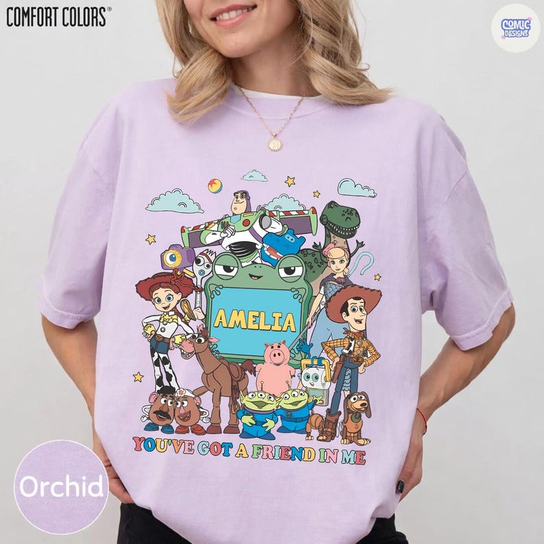 May include: A light purple short-sleeve t-shirt featuring a colorful graphic of various animated toy characters surrounding a green frog-shaped frame with the name AMELIA printed inside and the text YOU'VE GOT A FRIEND IN ME below.
