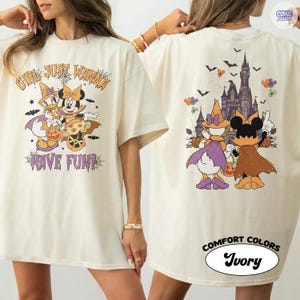 May include: An ivory t-shirt featuring a Halloween-themed graphic. The front displays Daisy Duck and Minnie Mouse with the text "GIRL JUST WANNA HAVE FUN!" The back shows the characters facing a castle with bats, balloons, and pumpkins.