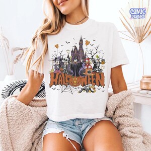 Puede incluir: Una camiseta blanca de manga corta con un gr&aacute;fico colorido de Halloween que muestra personajes de Disney disfrazados, un castillo oscuro y la palabra HALLOWEEN en letras grandes de color naranja, con murci&eacute;lagos y calabazas.