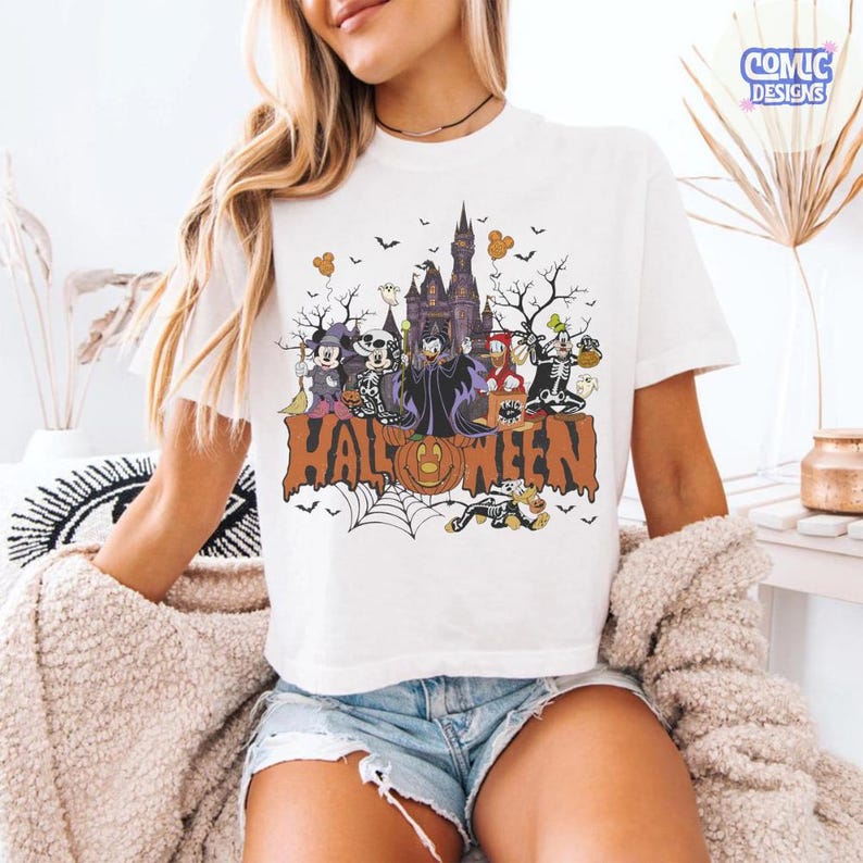 Puede incluir: Una camiseta blanca de manga corta con un gr&aacute;fico colorido de Halloween que muestra personajes de Disney disfrazados, un castillo oscuro y la palabra HALLOWEEN en letras grandes de color naranja, con murci&eacute;lagos y calabazas.