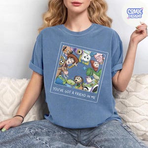 May include: A blue short-sleeve t-shirt featuring a square graphic print of various animated toy characters arranged in a circle. Below the graphic, the text reads, "YOU'VE GOT A FRIEND IN ME." The shirt has a crew neckline and a relaxed, casual fit.