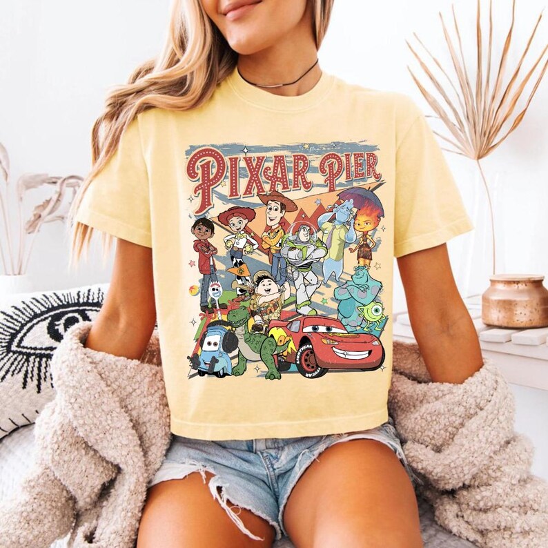 Pu&ograve; includere: Una maglietta giallo chiaro con una grafica colorata di vari personaggi Pixar, tra cui Woody, Buzz Lightyear e Saetta McQueen. Il testo "Pixar Pier" &egrave; visualizzato in uno stile vintage sopra i personaggi. La maglietta ha maniche corte.