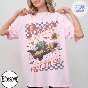 May include: A light pink t-shirt featuring a graphic print of a green three-eyed alien in a rocket ship. The design includes a checkered pattern, spider webs, pumpkins, and the text Pizza H-O-T-T-O-G-O and YOU CAN TAKE ME HOT TO GO.