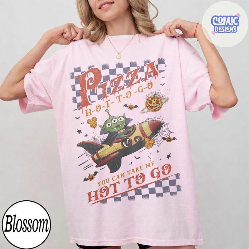 May include: A light pink t-shirt featuring a graphic print of a green three-eyed alien in a rocket ship. The design includes a checkered pattern, spider webs, pumpkins, and the text Pizza H-O-T-T-O-G-O and YOU CAN TAKE ME HOT TO GO.
