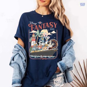 Puede incluir: Camiseta azul marino con un dise&ntilde;o de personajes de Disney en un crucero, con la inscripci&oacute;n "Disney Fantasy 2026". El dise&ntilde;o incluye un faro, palmeras y un tim&oacute;n. La palabra "NAME" tambi&eacute;n es visible.