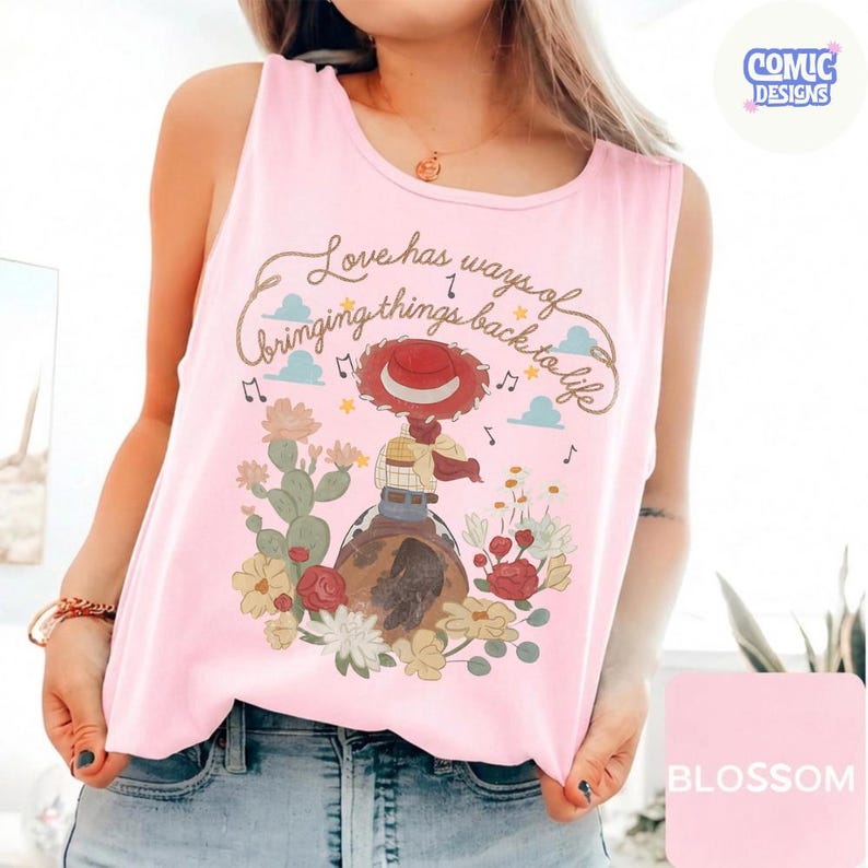 Op de afbeelding: Een lichtroze mouwloze tanktop met een grafische print van een cowgirl met een rode hoed, zittend op een paard te midden van cactussen en bloemen. De tekst luidt, Love has ways of bringing things back to life, met muzieknoten en wolken.