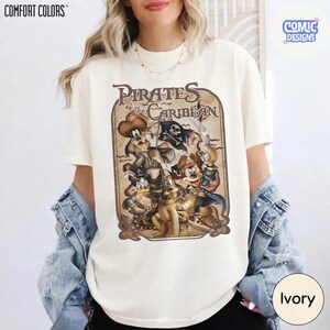 May include: Ivory-colored t-shirt featuring a vintage-style graphic of Disney characters as pirates, with the text "Pirates of the Caribbean." The design includes Mickey Mouse, Donald Duck, and Goofy. The shirt is from Comfort Colors and Comic Designs.
