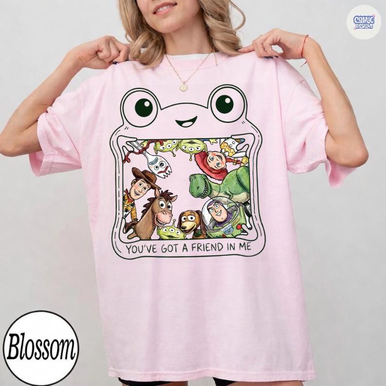 May include: A light pink t-shirt featuring a graphic of a frog-shaped frame containing various animated characters from the Toy Story franchise. The text "YOU'VE GOT A FRIEND IN ME" is printed in black capital letters below the character illustration.