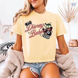 May include: A pale yellow t-shirt featuring the words "Disney Babe" in a cursive script, accompanied by Mickey Mouse ears, cherries, a billiard ball, and dice illustrations. The shirt has short sleeves.