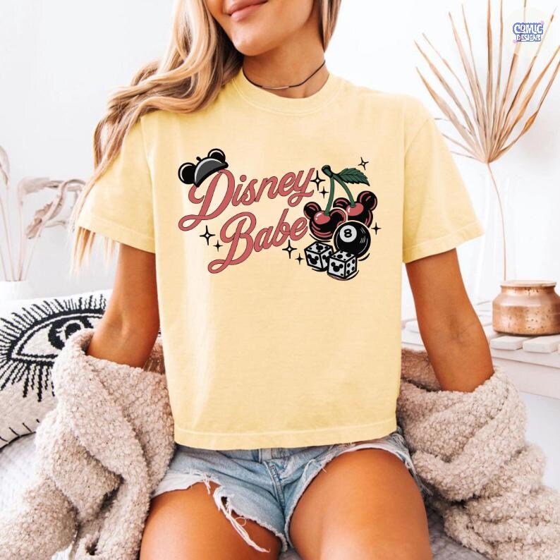 May include: A pale yellow t-shirt featuring the words "Disney Babe" in a cursive script, accompanied by Mickey Mouse ears, cherries, a billiard ball, and dice illustrations. The shirt has short sleeves.