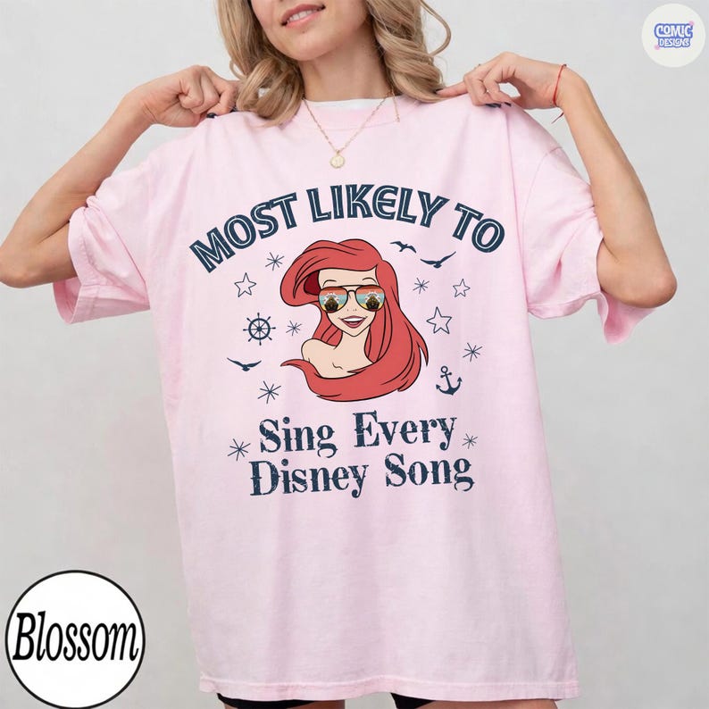 May include: Light pink t-shirt with the text "MOST LIKELY TO Sing Every Disney Song" in navy blue. The graphic features a cartoon of a red-haired character wearing sunglasses. Additional nautical-themed graphics are included.