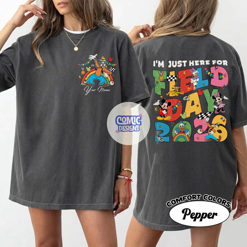K&ouml;nnte beinhalten: Dunkelgraues T-Shirt mit einer bunten Grafik auf der Vorder- und R&uuml;ckseite. Die Vorderseite zeigt eine Zeichentrickfigur &uuml;ber einem Regenbogen mit dem Text "Your Name". Auf der R&uuml;ckseite steht "I'M JUST HERE FOR FIELD DAY 2023" mit Zeichentrickfiguren.