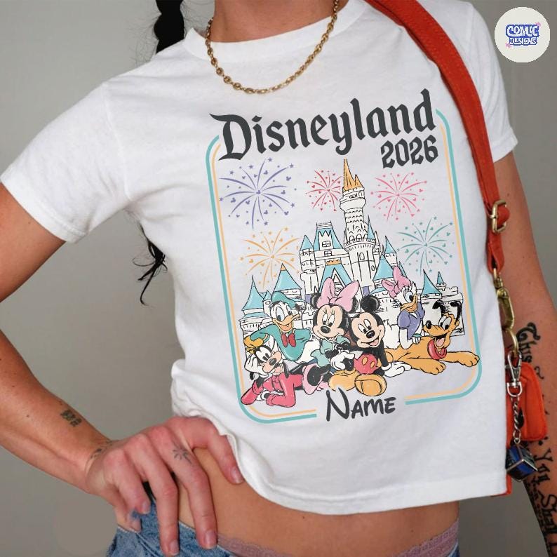 Op de afbeelding: Wit T-shirt met een kleurrijke Disneyland 2026 afbeelding. Het ontwerp bevat een kasteel, vuurwerk en Disney-personages zoals Mickey en Minnie Mouse, Donald Duck en Goofy. Het shirt heeft een ronde hals.
