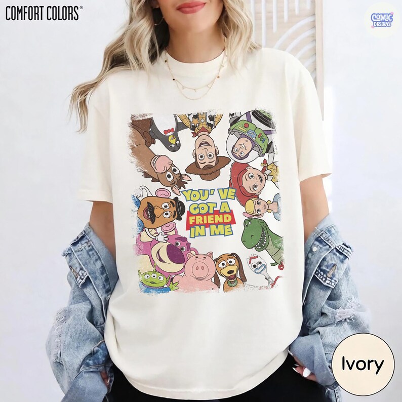 May include: Ivory-colored t-shirt featuring a colorful graphic of characters from the animated film "Toy Story." The design includes Woody, Buzz Lightyear, and other characters, with the phrase "You've Got a Friend in Me."