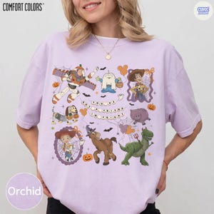 May include: A light purple t-shirt featuring a graphic print of various animated characters in Halloween costumes, including a space ranger, a cowboy, and a dinosaur. The text "YOU'VE GOT A FRIEND IN FRIGHT" is centered among spiderwebs, pumpkins, and bats.