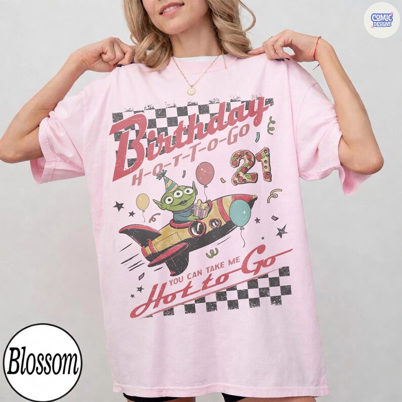 May include: A light pink t-shirt with a graphic featuring a cartoon alien in a rocket ship, balloons, and the text "Birthday H-O-T-T-O-GO 21". The shirt has a checkerboard design and the text "You can take me Hot to Go".