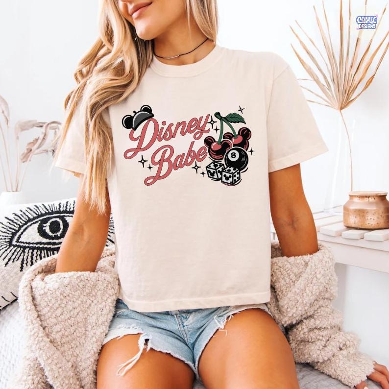 May include: A cream-coloured t-shirt featuring the text "Disney Babe" in pink script, alongside a graphic of cherries, an eight ball, and dice. The shirt has short sleeves and a crew neck.