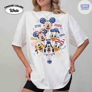 以下が含まれることがあります： 白のComfort Colors Tシャツ。ミッキーマウスとミニーマウス、ドナルドダックとデイジーダック、グーフィーとプルートなど、ディズニーのキャラクターが風船になったグラフィック。愛国的な色と「1776 2026 USA 250th」の文字がデザインされています。