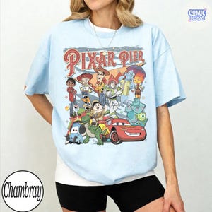 Pu&ograve; includere: T-shirt azzurra con una grafica colorata dei personaggi di Pixar Pier. Il design include personaggi di Toy Story, Monsters, Inc., Cars e altro ancora. Il testo "Pixar Pier" &egrave; visualizzato in un font retr&ograve;. La maglietta ha maniche corte.