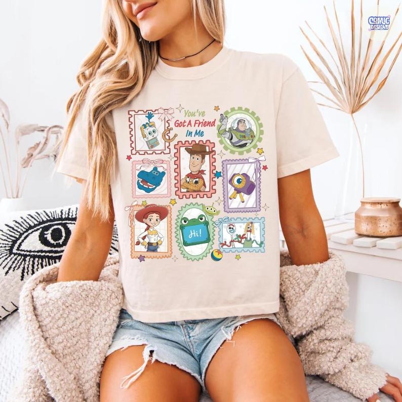 May include: A cream-colored t-shirt featuring a graphic print of various Toy Story characters inside colorful postage stamp-style frames. The text "You've Got A Friend In Me" is printed in the center. The design includes Woody, Buzz Lightyear, Jessie, Rex, and other characters with small stars and decorative elements.