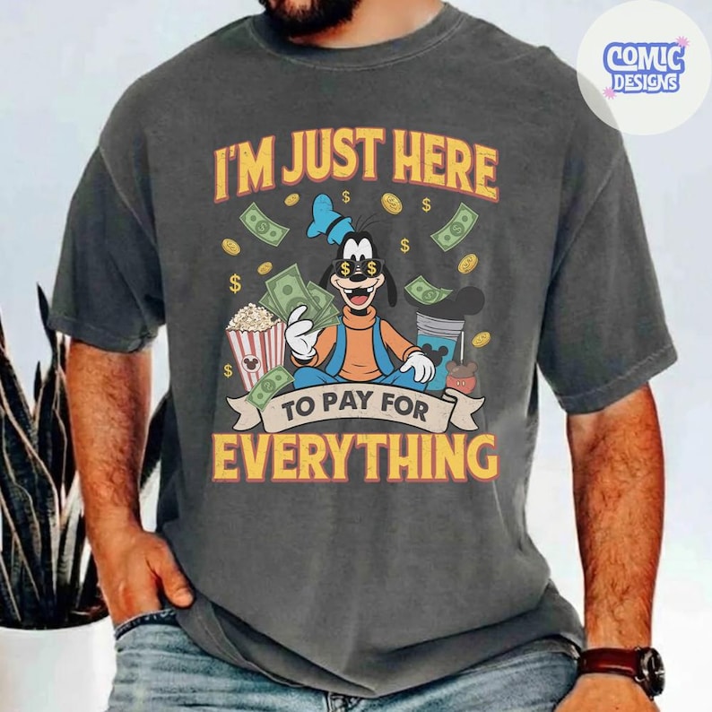 May include: Dark gray t-shirt featuring a cartoon image of Goofy with the text "I'M JUST HERE TO PAY FOR EVERYTHING." The design includes dollar bills, popcorn, and other elements, with a Comic Designs logo in the upper right corner.