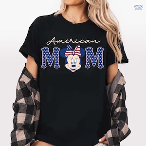 May include: Black t-shirt with the words "American MOM" in white script and blue block letters. The "MOM" letters are designed with a pattern of blue stars on a white background. A Minnie Mouse graphic with a patriotic bow is in the center.
