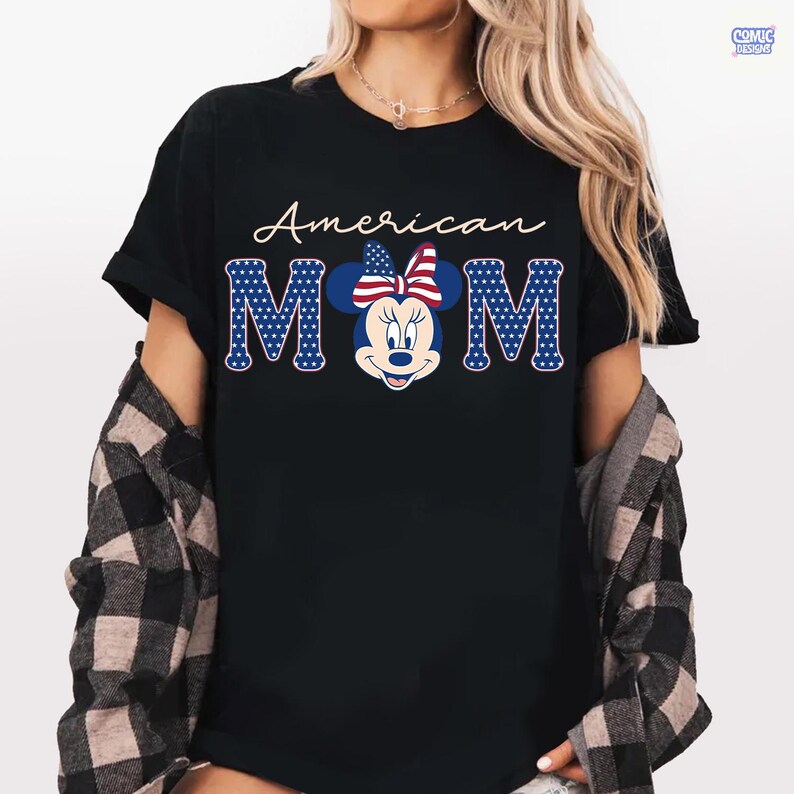 May include: Black t-shirt with the words "American MOM" in white script and blue block letters. The "MOM" letters are designed with a pattern of blue stars on a white background. A Minnie Mouse graphic with a patriotic bow is in the center.