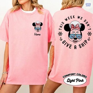 May include: Light pink t-shirt featuring a Minnie Mouse graphic on the front and back. The front has a Minnie Mouse face with sunglasses and the word "Name". The back says "This week we don't give a ship" with Minnie Mouse and nautical elements.