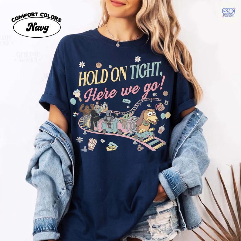 May include: Navy blue t-shirt with the text "HOLD ON TIGHT, Here we go!" and a cartoon roller coaster design featuring characters. The shirt has the "COMFORT COLORS" and "COMIC DESIGNS" logos.