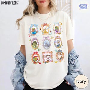 Puede incluir: Camiseta Comfort Colors color marfil con personajes de dibujos animados enmarcados. Los marcos dorados tienen lazos coloridos y detalles florales. El logotipo de Comic Designs est&aacute; en la esquina superior derecha. La camiseta es marfil.